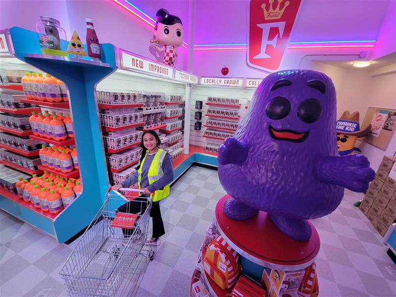 Funmart: A Funko volunteer pushes a grocery cart past aisles of 3-liter SODAs and other collectibles, while a Grimace statue balances on top of a McDonald’s Loungefly display.